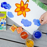 An overhead view of the Natural Earth Paint Petite Paint Kit in use. A child's hand can be seen holding the paintbrush, painting blue clouds.