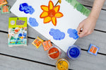 An overhead view of the Natural Earth Paint Petite Paint Kit in use. A child's hand can be seen holding the paintbrush, painting blue clouds.