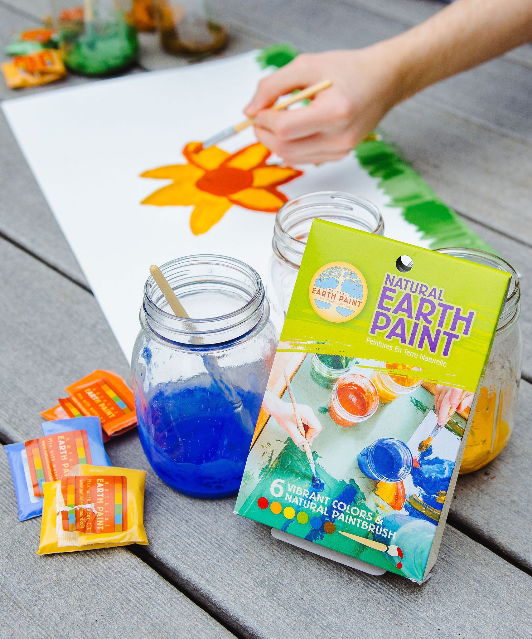 The Natural Earth Paint Petite Paint Kit in use. A child's hand can be seen in the background painting a picture of a daffodil.