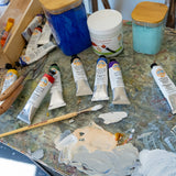 Natural Earth Paint acrylic aluminium paint tubes on a wooden surface in use to mix paint with the eco wooden brush, the palette knife and acrylic medium