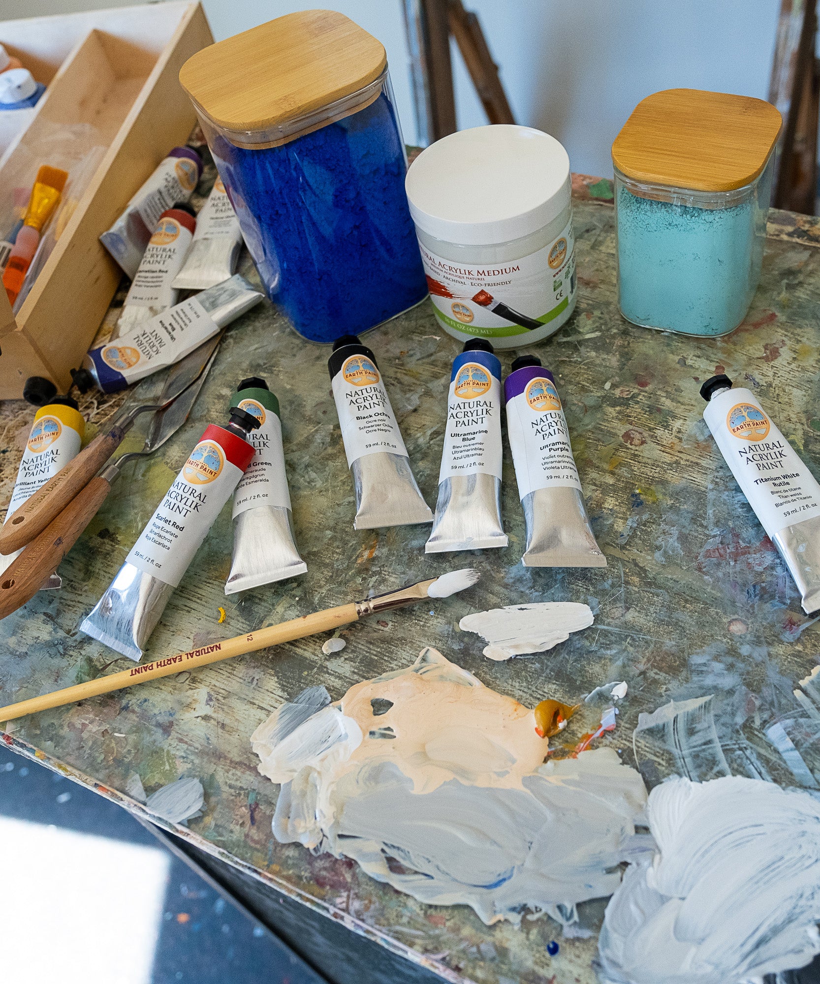Natural Earth Paint acrylic aluminium paint tubes on a wooden surface in use to mix paint with the eco wooden brush, the palette knife and acrylic medium