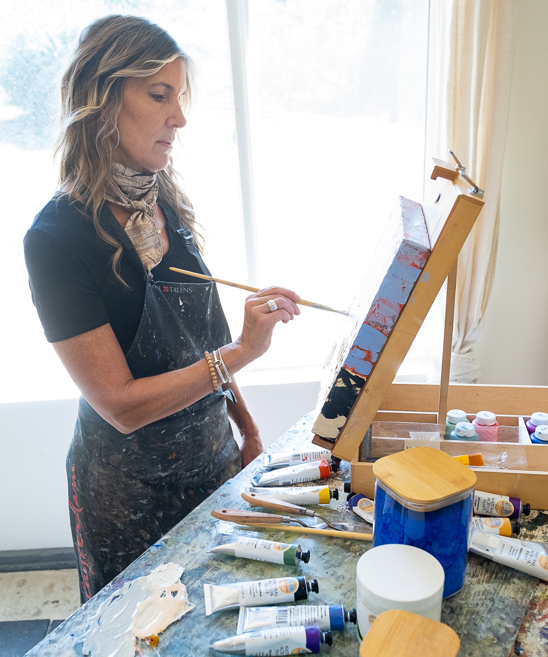 An artist in the studio painting on a canvas with the Natural Earth Pain natural acrylic paints resting on a wooden surface