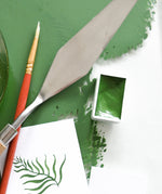 Natural Earth Paint watercolour pan filled with green paint next to a palette knife and brush