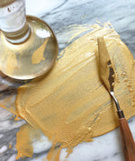 Natural Earth Paint palette knife with wooden handle spreading gold glittery paint on a stone surface.