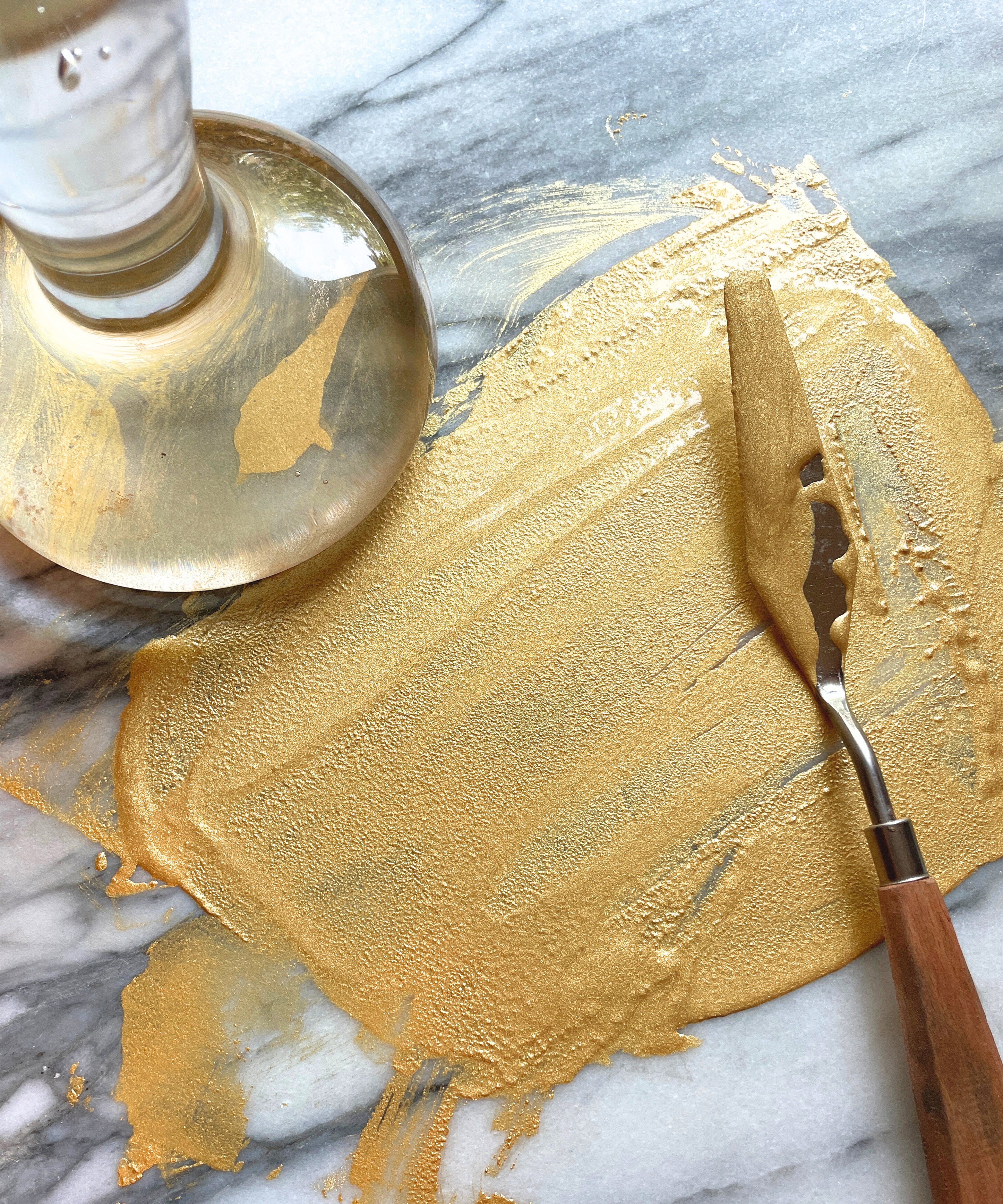 Natural Earth Paint palette knife with wooden handle spreading gold glittery paint on a stone surface.