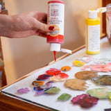 A close up of a hand squeezing the red Natural Earth Paint acrylic paint from the bottle onto a glass surface showing the constiency