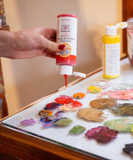 A close up of a hand squeezing the red Natural Earth Paint acrylic paint from the bottle onto a glass surface showing the constiency