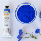 A top down view of the Natural earth paint in ultramarine blue next to the blue pigment powder in a glass bowl next to blue flowers