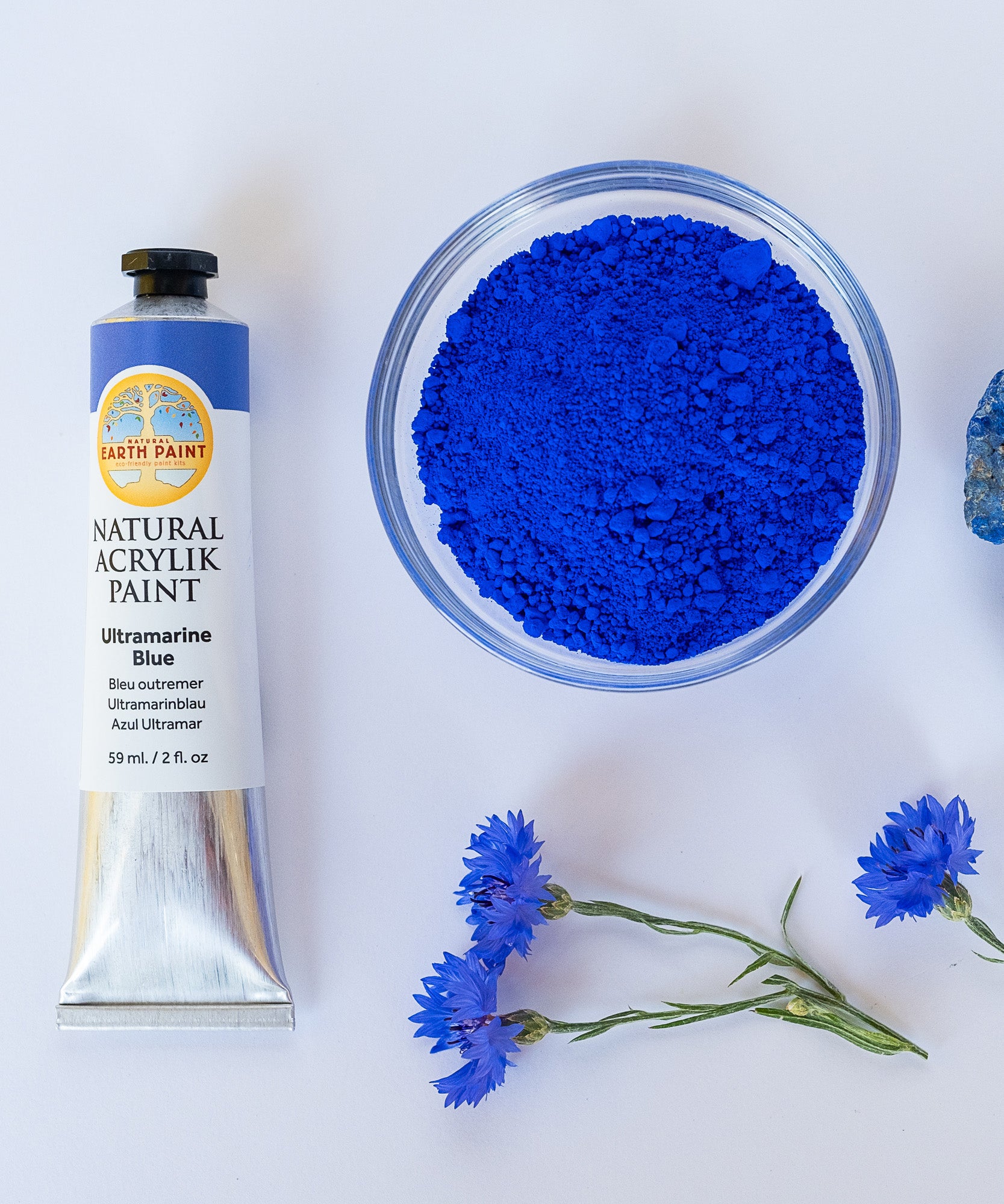 A top down view of the Natural earth paint in ultramarine blue next to the blue pigment powder in a glass bowl next to blue flowers