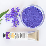 A top down view of the Natural earth paint tube in ultramarine purple next to a glass  bowl of matching paint pigment and a bunch of purple flowers