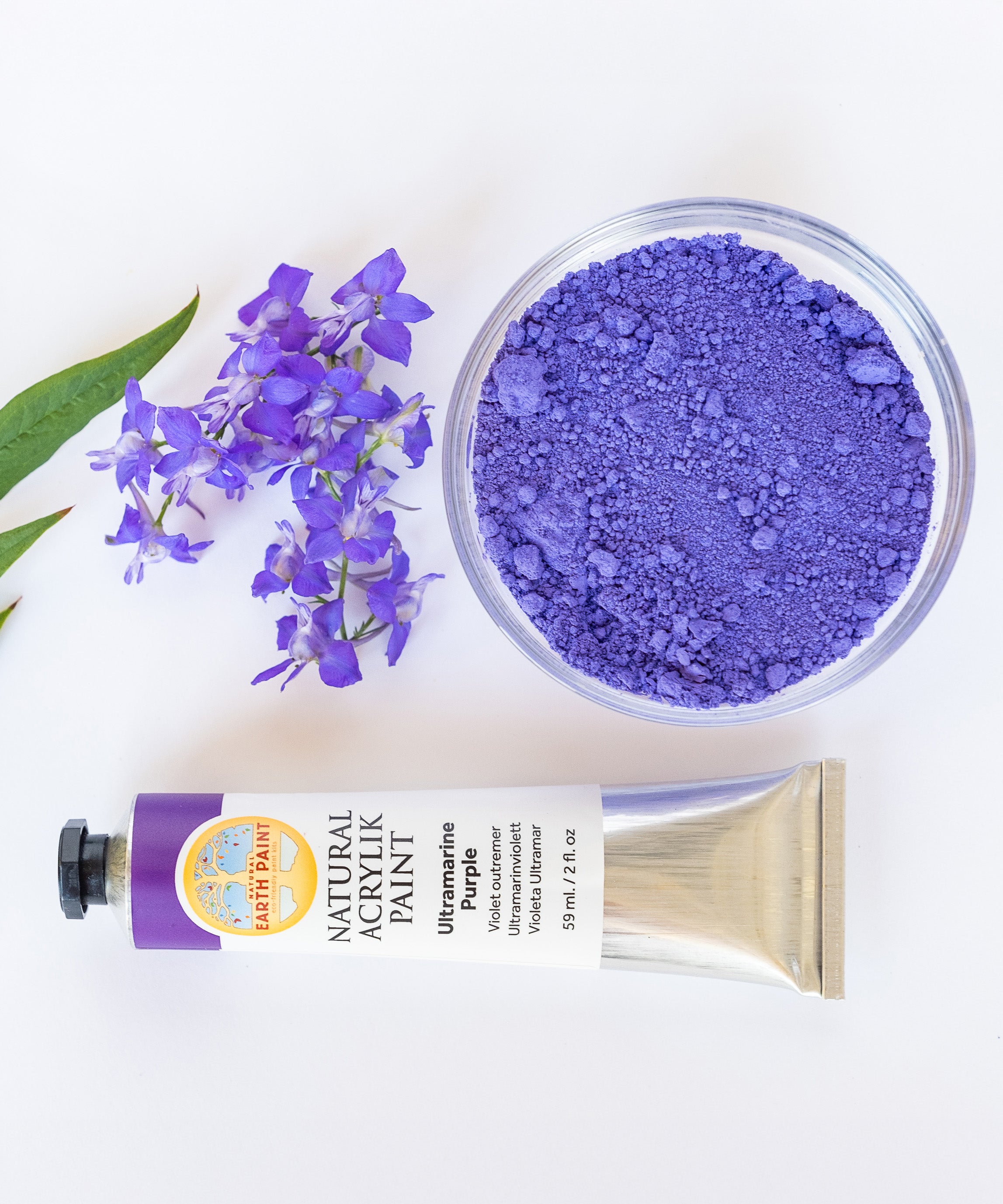 A top down view of the Natural earth paint tube in ultramarine purple next to a glass  bowl of matching paint pigment and a bunch of purple flowers