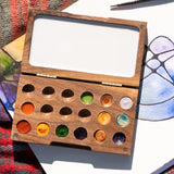 A close up of the Natural Earth Paint small wooden watercolour palette on top of a canvas showing different coloured paint in the well and a blank mixing panel