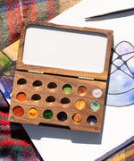 A close up of the Natural Earth Paint small wooden watercolour palette on top of a canvas showing different coloured paint in the well and a blank mixing panel