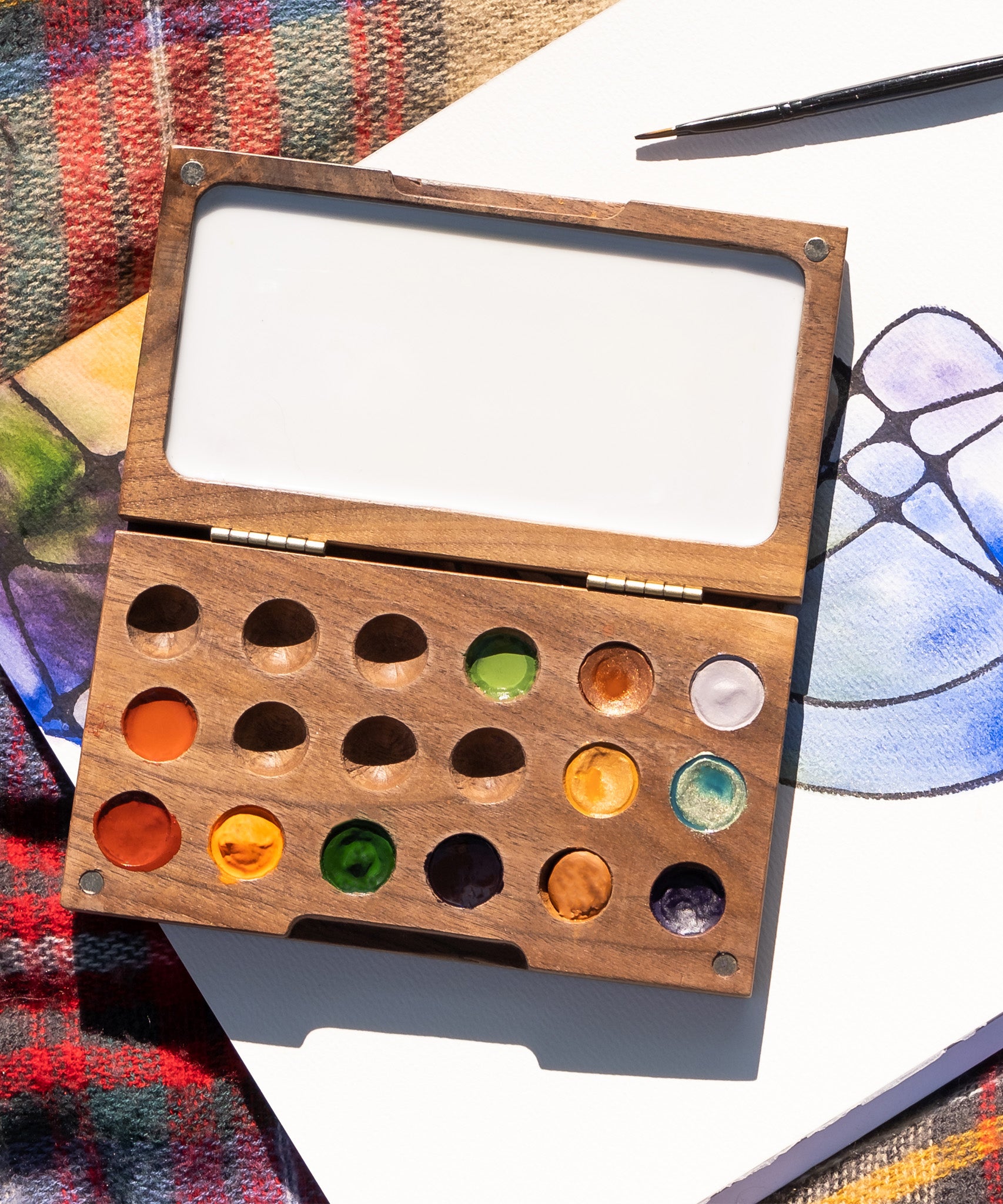 A close up of the Natural Earth Paint small wooden watercolour palette on top of a canvas showing different coloured paint in the well and a blank mixing panel
