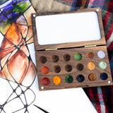 The  Natural Earth Paint small wooden watercolour palette on top of a canvas with a abstract painting showing different coloured paint in the well and little magnetic pieces on each corner to open and close