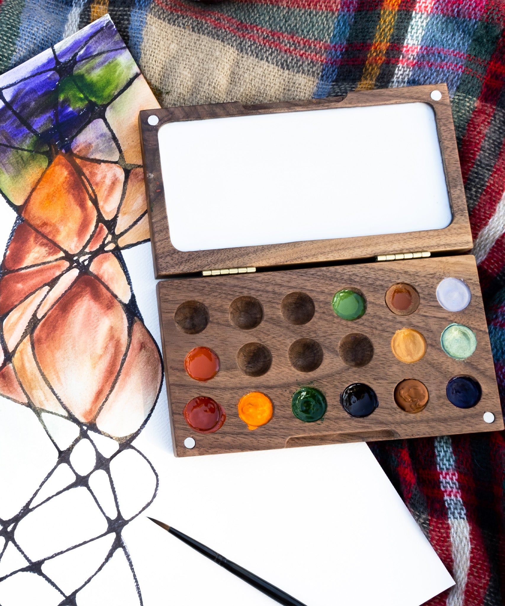 The  Natural Earth Paint small wooden watercolour palette on top of a canvas with a abstract painting showing different coloured paint in the well and little magnetic pieces on each corner to open and close