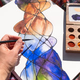 A close up of the Natural Earth Paint small wooden watercolour palette on top of a canvas with an adult hand holding a paint brush showing the scale of the palette.