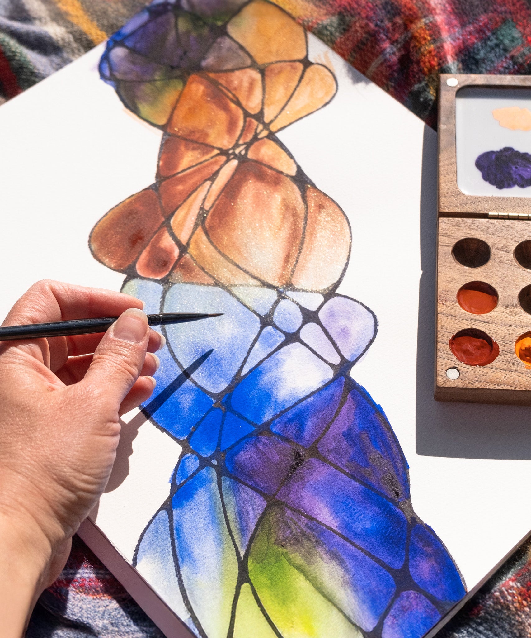 A close up of the Natural Earth Paint small wooden watercolour palette on top of a canvas with an adult hand holding a paint brush showing the scale of the palette.