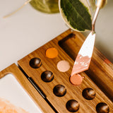 A close up view of the Natural Earth Paint wooden palette showing the palette knife scooping paint from the hole