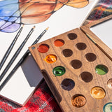 A close up of the Natural Earth Paint small wooden watercolour palette on top of a canvas showing the paint well detail with different coloured paint inside next to paint brushes