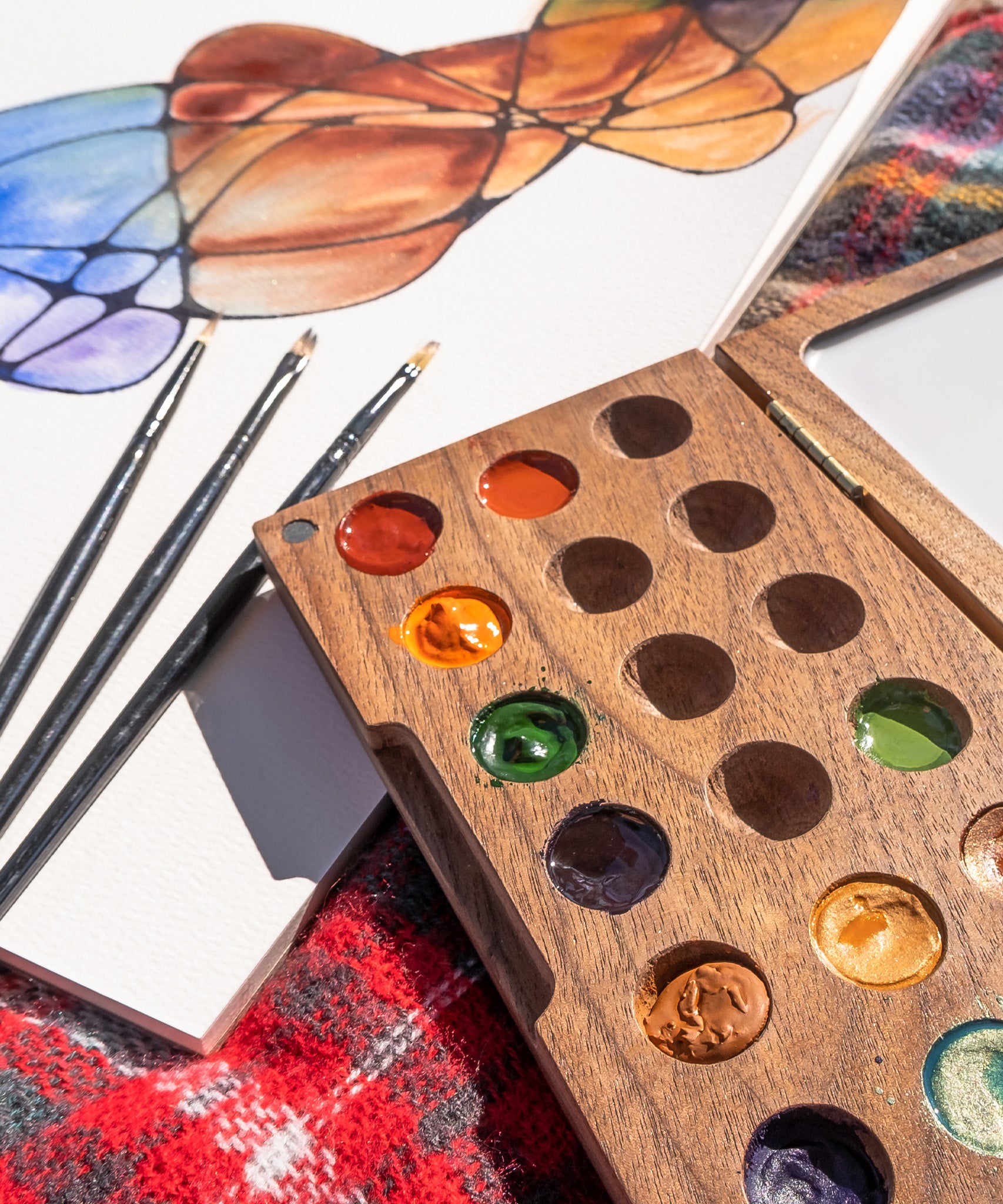 A close up of the Natural Earth Paint small wooden watercolour palette on top of a canvas showing the paint well detail with different coloured paint inside next to paint brushes