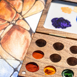 A close up of the Natural Earth Paint small wooden watercolour palette on top of a canvas showing different coloured paint in the wells and mixed paint on the white mixing panel