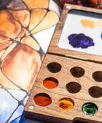 A close up of the Natural Earth Paint small wooden watercolour palette on top of a canvas showing different coloured paint in the wells and mixed paint on the white mixing panel