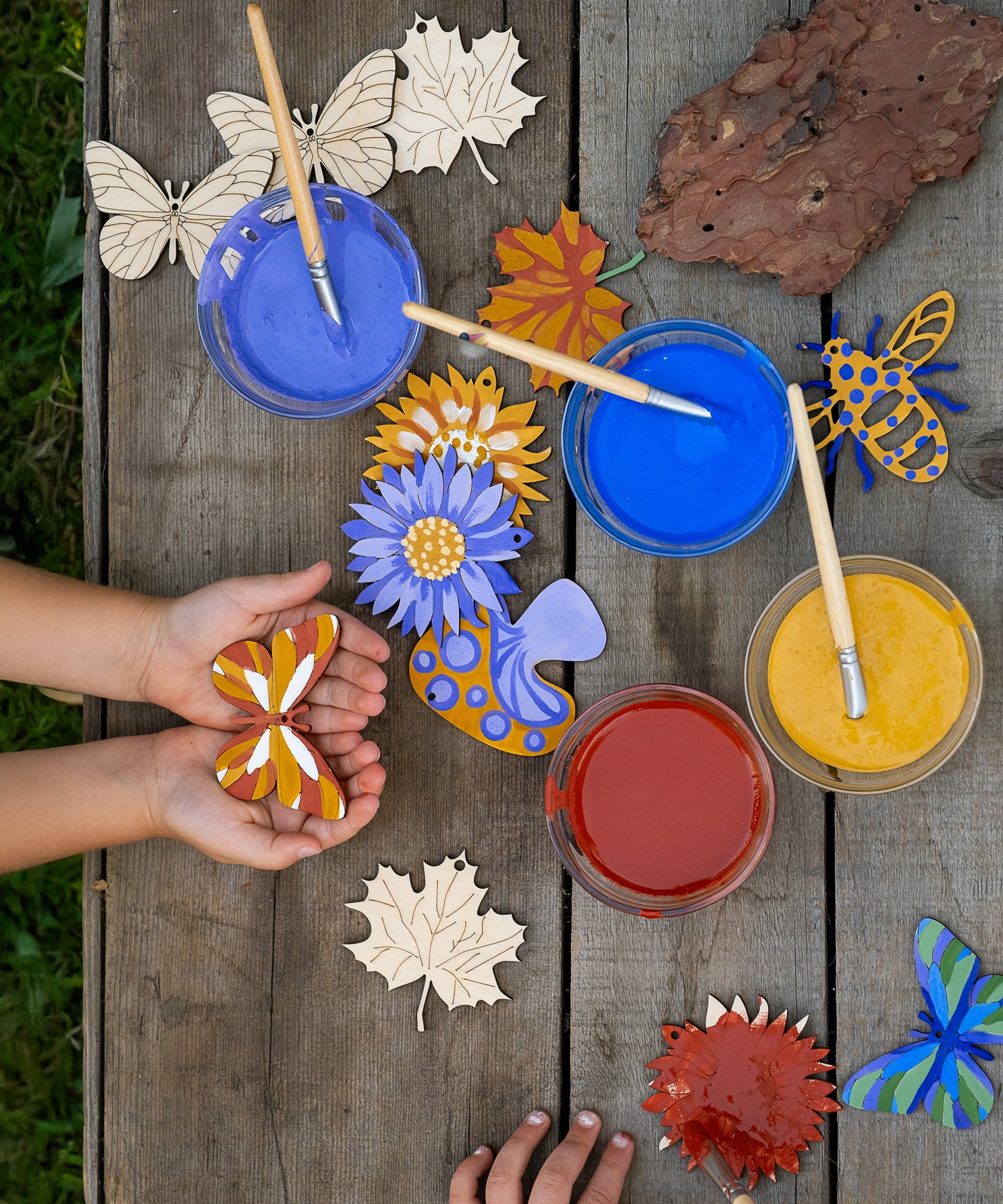 An overhead view of children using the Natural Earth Paint nature craft kit. they can be seen painting and holding their painted wooden decorations