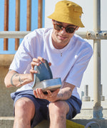 A man sitting on a stone surface and lifting the pale blue silicone lid from the soldier One Green Bottle lunchbox.