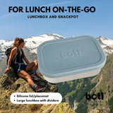 A One Green Bottle lunchbox in soldier colour with a mountain range in the background. A stainless steel plastic free lunchbox available at Bapipur. Features a pale blue silicone lid, perfect for lunch on the go