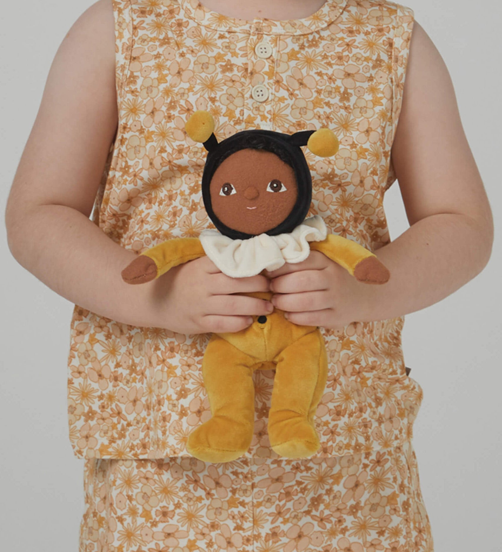 A little girl holding the Olli Ella Dinky Dinkum Doll - Billie Bumblebee - in a yellow onesie with a black hood and antennae, with a white ruffled collar, brown eyes, and buzzy bee wings.