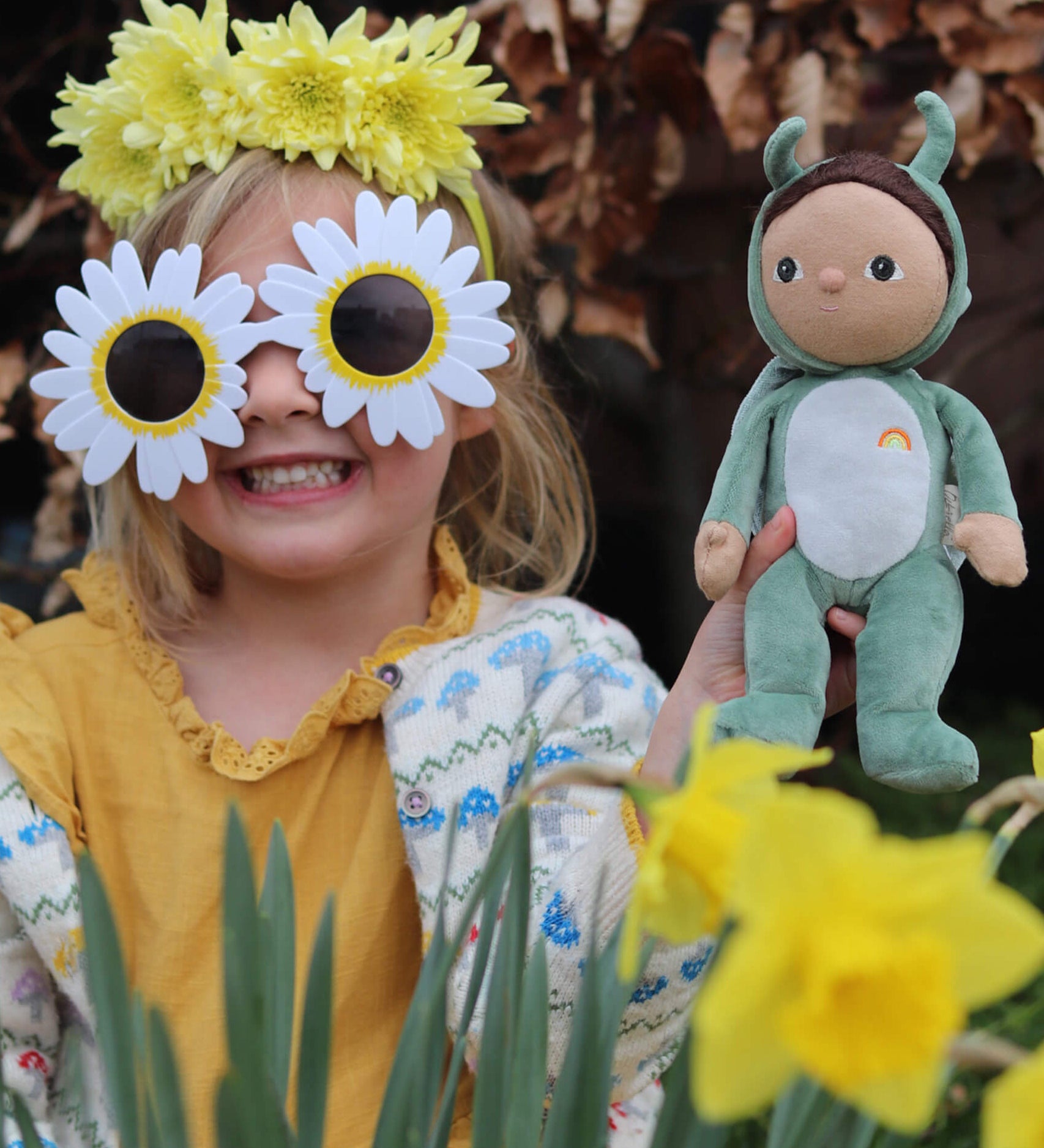 A little girl in flower sunglasses holding the Olli Ella Dinky Dinkum Doll - Gideon Grasshopper - in a garden.