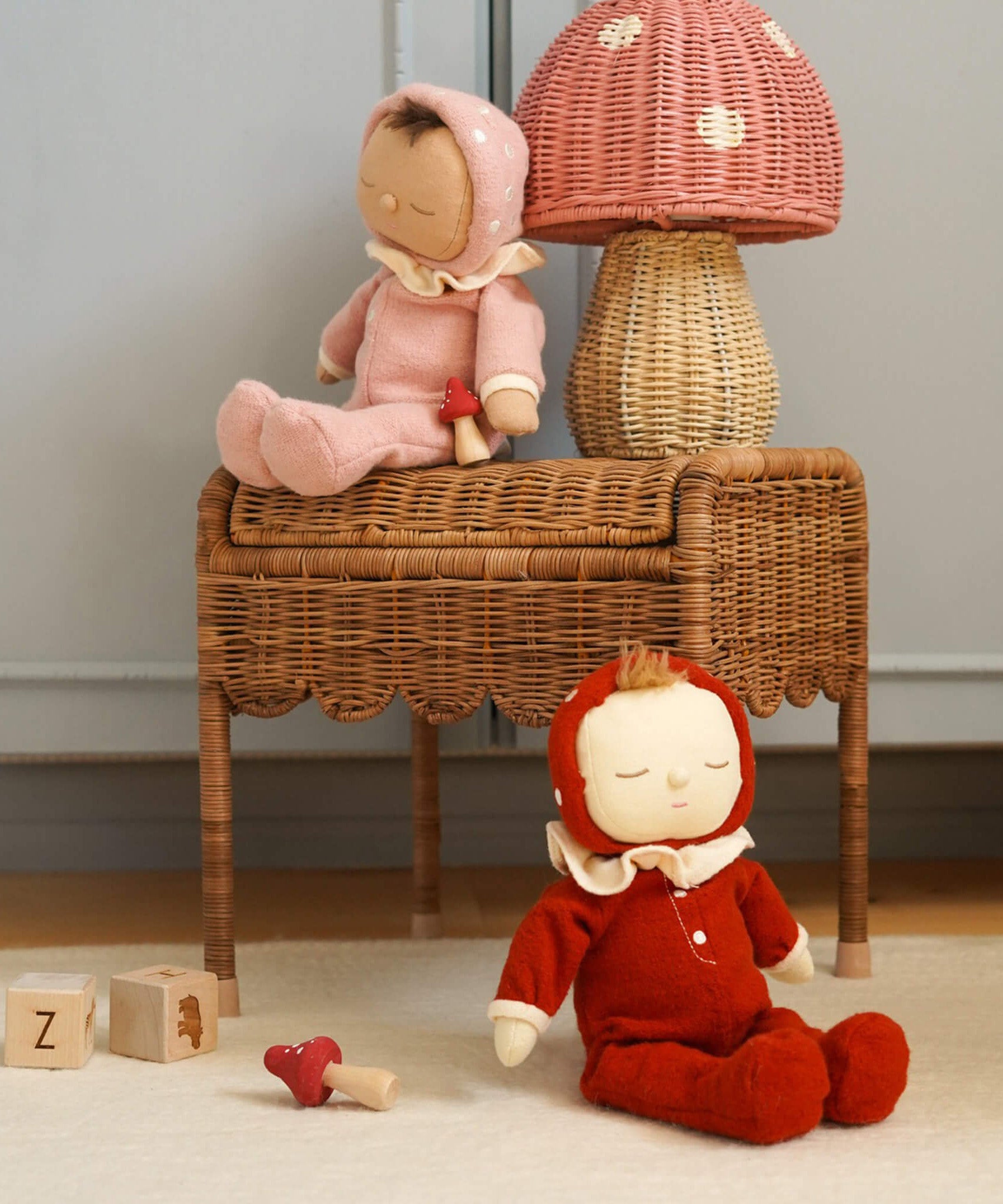 The Olli Ella rattan toadstool lamp with a natural base and a pink mushroom top, on a table with dinkum dolls.
