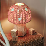 The Olli Ella rattan toadstool lamp with a natural base and a pink mushroom top, projecting soft warm light in a bedroom.