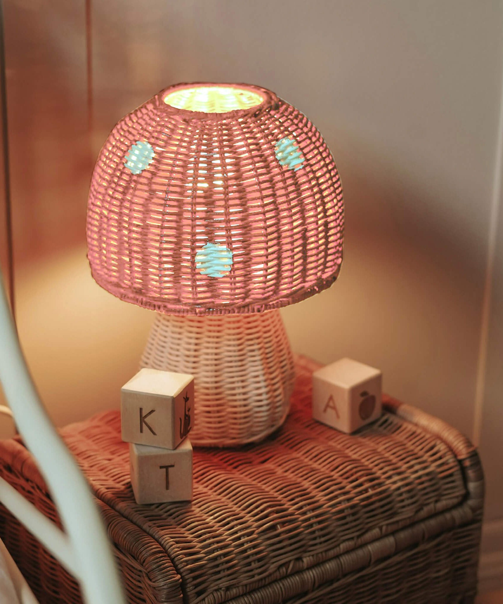 The Olli Ella rattan toadstool lamp with a natural base and a pink mushroom top, projecting soft warm light in a bedroom.