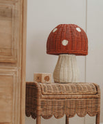 The Olli Ella rattan toadstool lamp with a white base and a red mushroom top, on a rattan table.