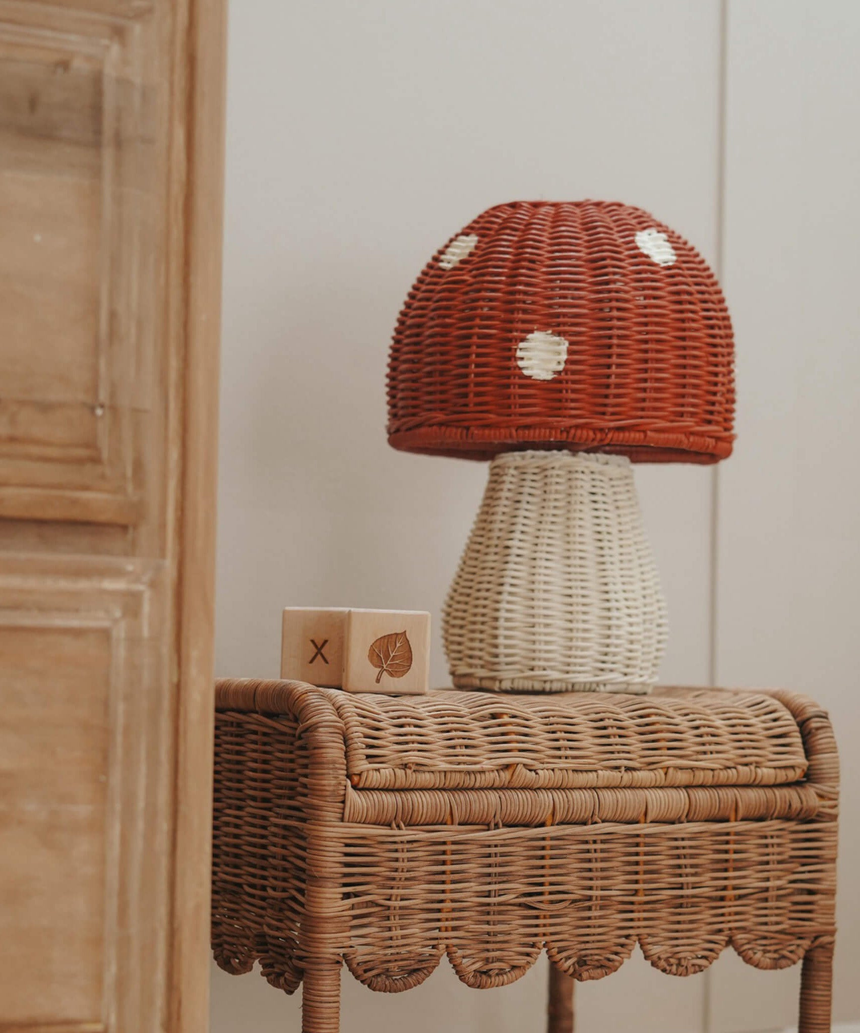 The Olli Ella rattan toadstool lamp with a white base and a red mushroom top, on a rattan table.