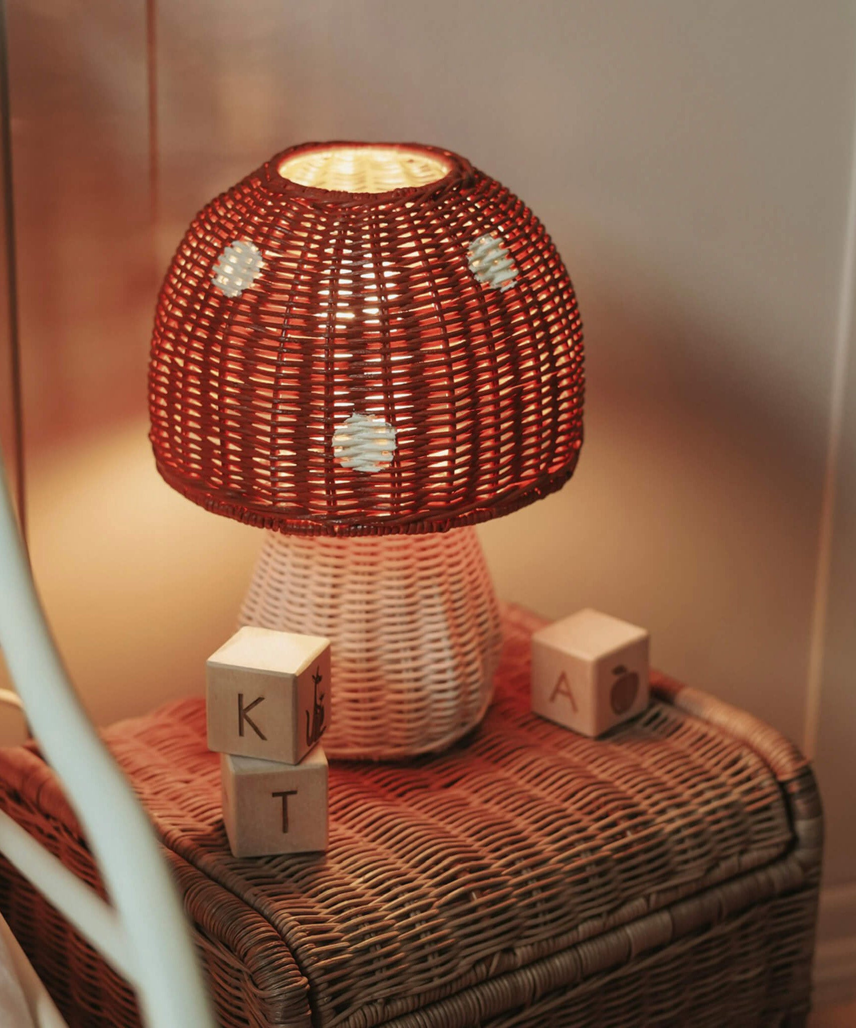 The Olli Ella rattan toadstool lamp with a white base and a red mushroom top, on a toy box and creating oft warm light.