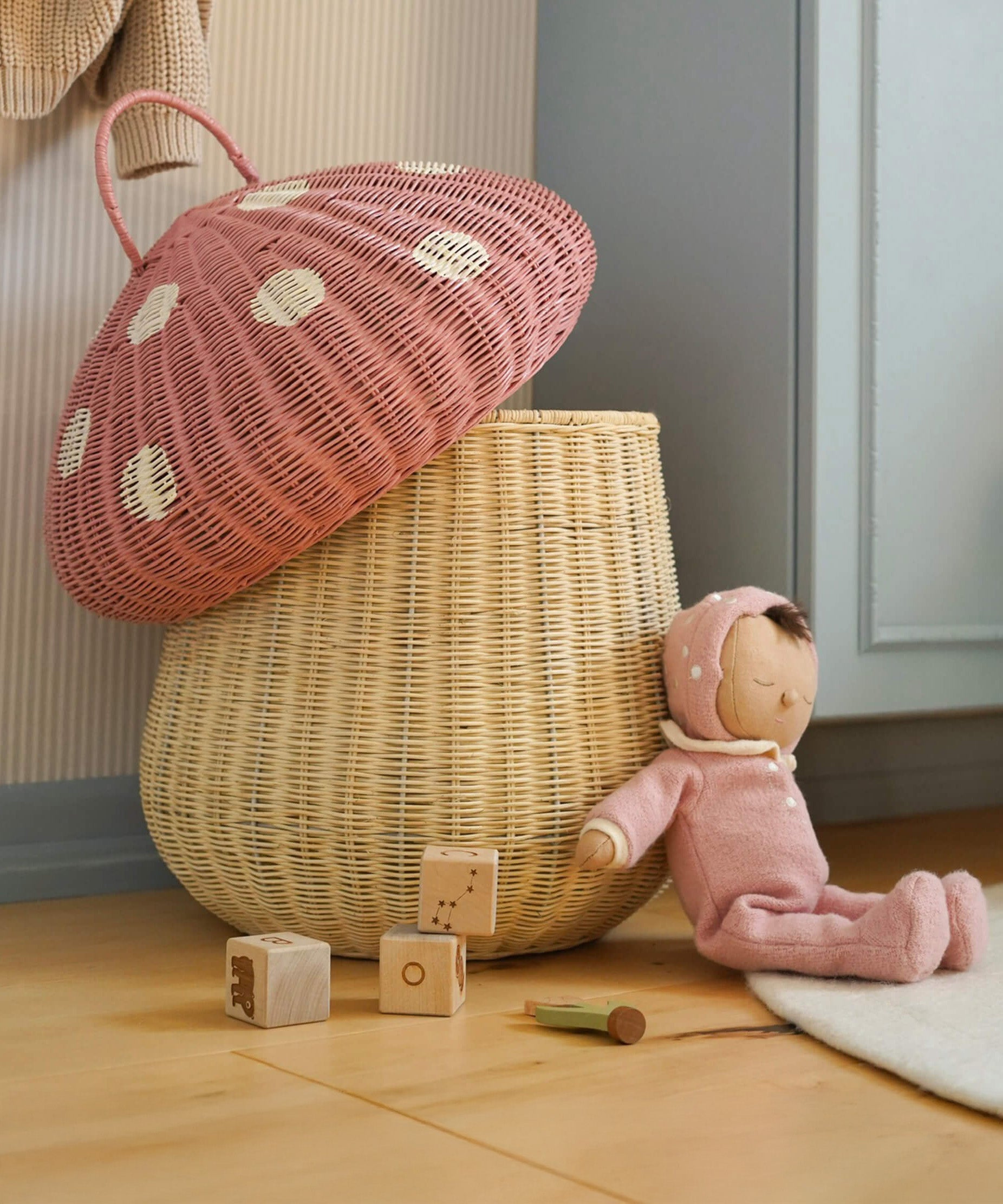 The Olli Ella large woven rattan storage basket, in the shape of a mushroom with a spotty pink toadstool lid, a dozy doll is leaning against it.