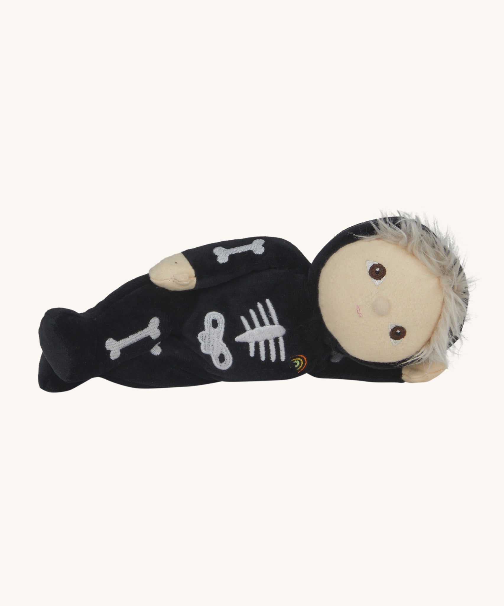 Olli Ella Scully Skeleton Dinky Dinkum Doll soft toy laying on it's side. The doll features glow in the dark bone details on the front along with a small embroidered rainbow on the chest. These soft toys are part of Olli Ella's Halloween collection available here at Babipur.