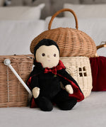 An Olli Ella Vincent Vampire Dinky Dinkum doll placed in a seated position in front of baskets. A soft vampire doll with a black cape with bat shaped glow in the dark details on the back. These soft toys are part of Olli Ella's Halloween collection available here at Babipur.