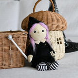 An Olli Ella Willa Witch Dinky Dinkum Doll placed in a seated position in front of baskets. A witch with a black outfit and matching hat with glow in the dark features on the striped tights, collar and stars on the hat. These soft toys are part of Olli Ella's Halloween collection available here at Babipur.