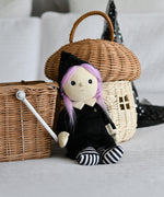 An Olli Ella Willa Witch Dinky Dinkum Doll placed in a seated position in front of baskets. A witch with a black outfit and matching hat with glow in the dark features on the striped tights, collar and stars on the hat. These soft toys are part of Olli Ella's Halloween collection available here at Babipur.
