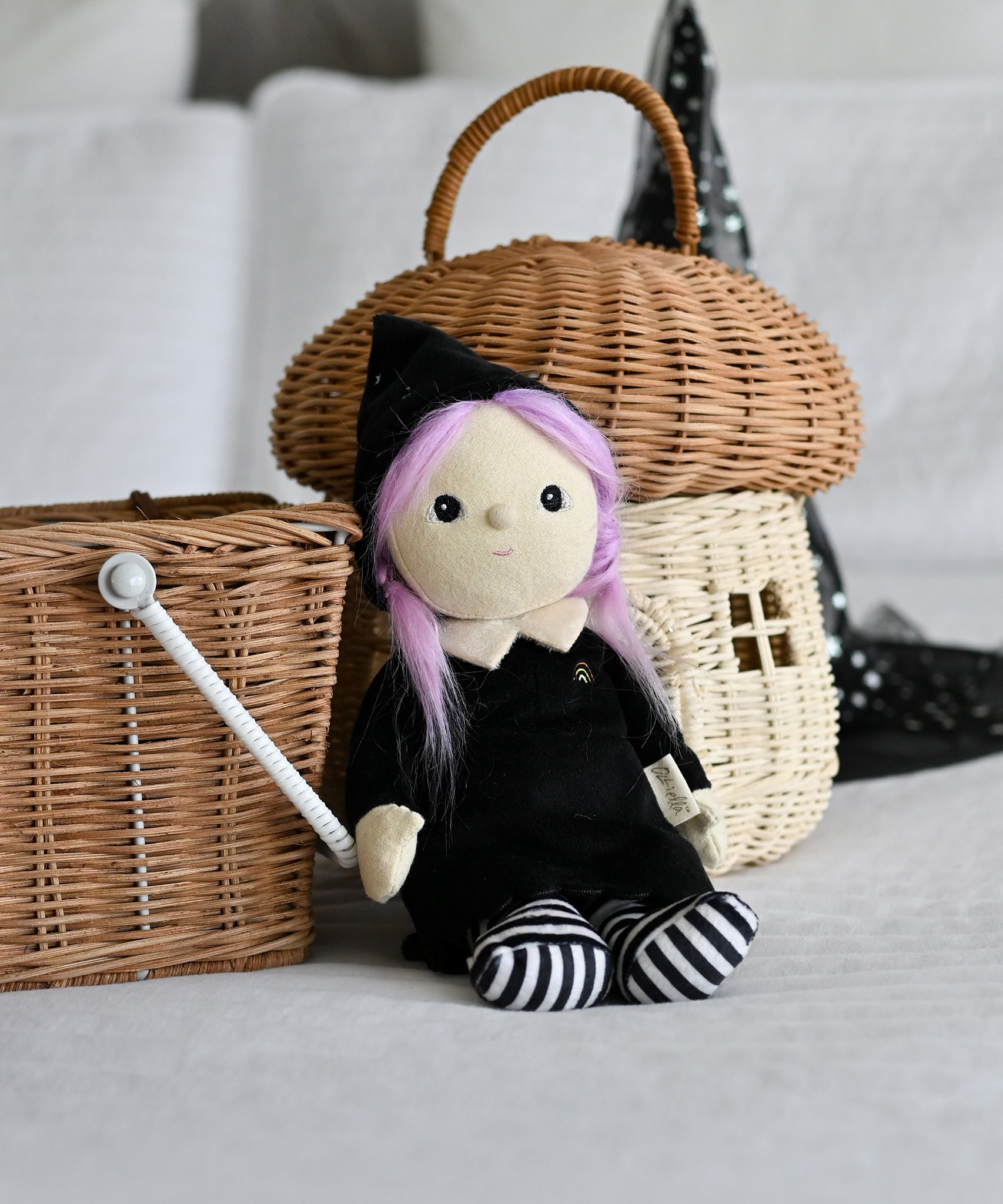 An Olli Ella Willa Witch Dinky Dinkum Doll placed in a seated position in front of baskets. A witch with a black outfit and matching hat with glow in the dark features on the striped tights, collar and stars on the hat. These soft toys are part of Olli Ella's Halloween collection available here at Babipur.
