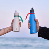 Two hands holding One Green Bottle canteens with Sloop bottle holders around the next in front of a sun set