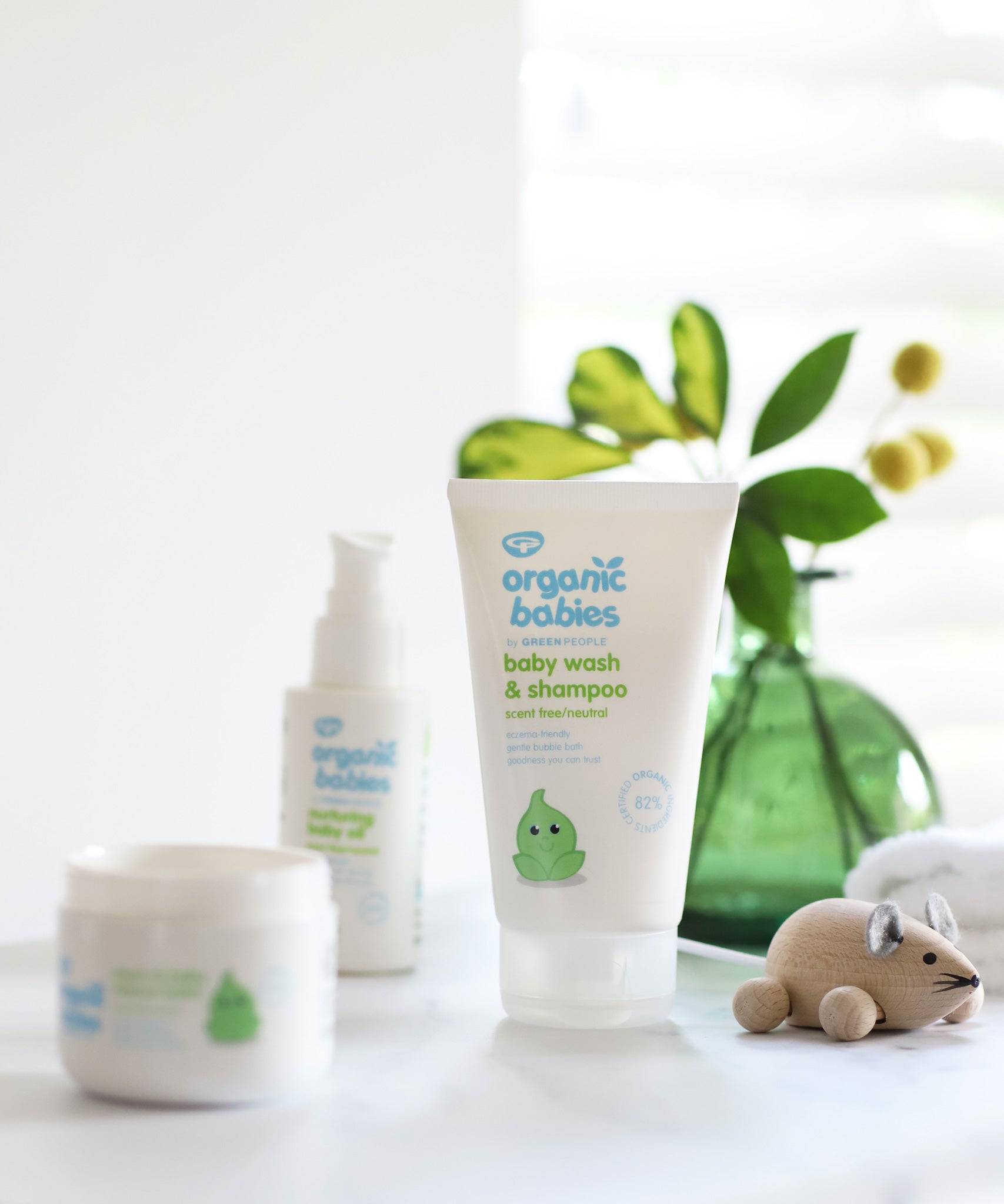 Organic Babies Baby Wash & Shampoo Scent Free placed on a white surface next to a wooden toy mouse. Other products from the organic babies collection can be seen in the background.
