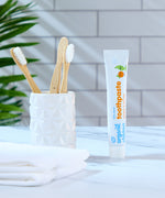 A tube of Organic Babies Toothpaste Mandarin & Aloe Vera pictured next to a pot of bamboo toothbrushes. A green leafy plant can be seen in the background.
