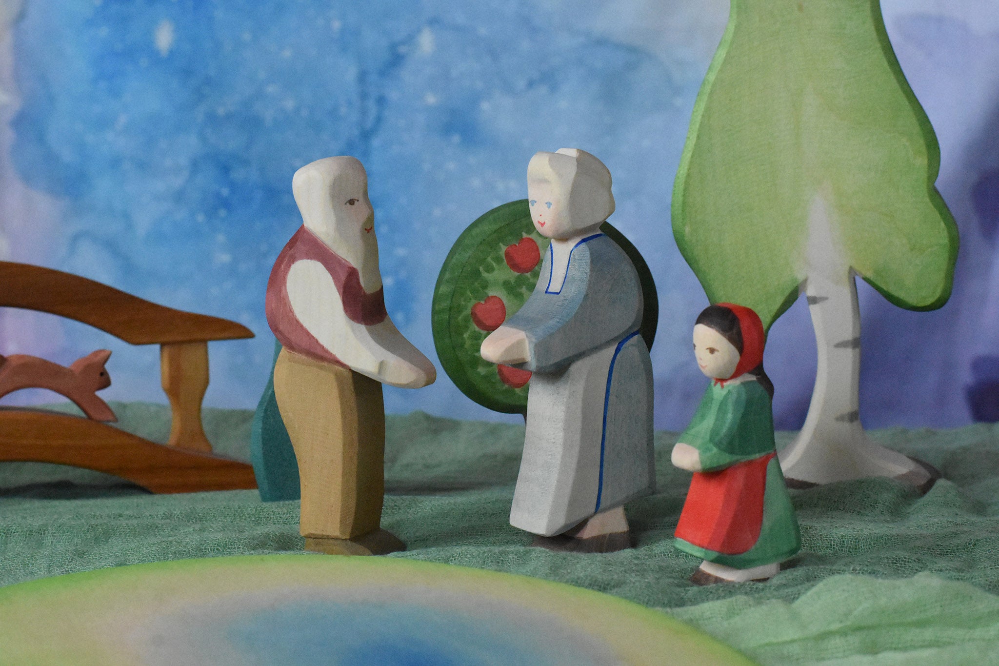 An Ostheimer grandmother, grandfather and little red riding hood figure placed in a small world play scene. There is a green coloured decorative cloth covering the surface beneath them and an apple tree and large birch tree in the background.  These wooden figures are part of a wide range of expertly hand crafted wooden toys here at Babipur.