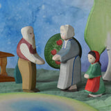 An Ostheimer grandmother, grandfather and little red riding hood figure placed in a small world play scene. There is a green coloured decorative cloth covering the surface beneath them and an apple tree and large birch tree in the background.  These wooden figures are part of a wide range of expertly hand crafted wooden toys here at Babipur.