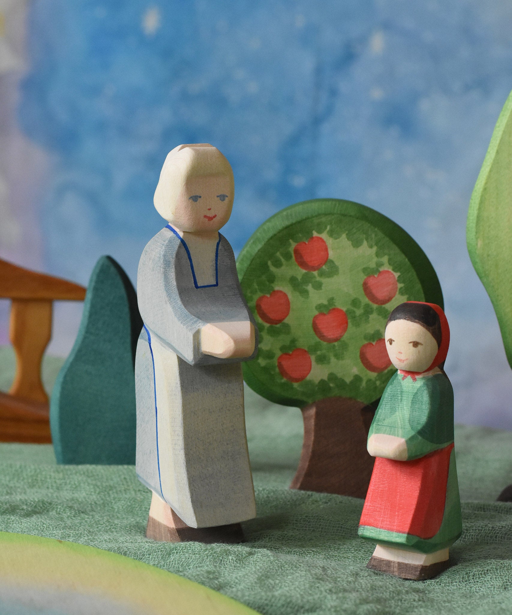 An Ostheimer grandmother figure and little red riding hood figure placed in a small world play scene. There is a green coloured decorative cloth covering the surface beneath them and an apple tree and green bush in the background.  These wooden figures are part of a wide range of expertly hand crafted wooden toys here at Babipur.